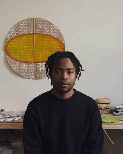 headshot of Kern Samuel in his studio, wearing a dark shirt, posed in front of paper artwork on the wall