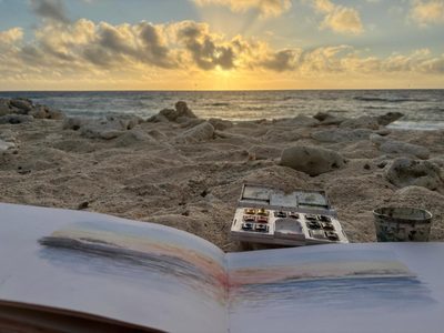 Open sketchbook and watercolors on a beach with sunset and clouds in the background