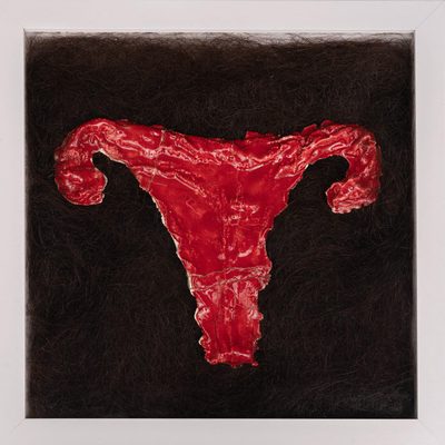 Red, ceramic-glazed underwear, simultaneously resembling a ram's head/horns and a womb/ovaries. The ceramic piece is resting within a square of dark human hair.
