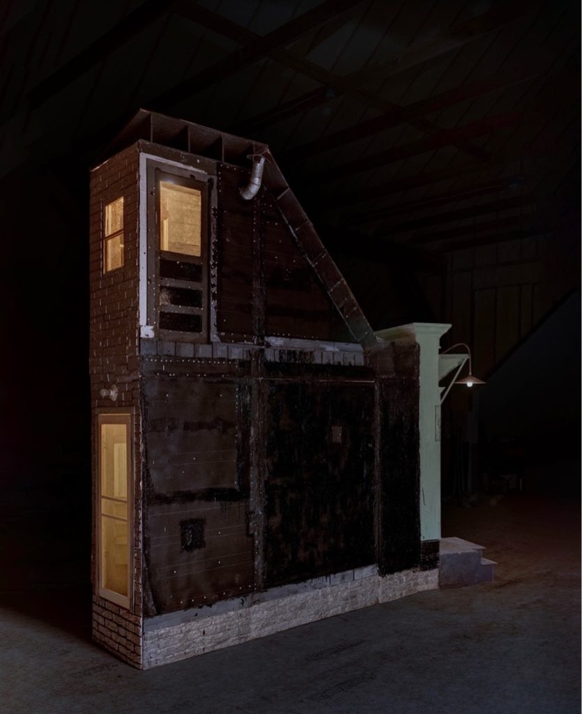 A dark photo of a large sculpture. The sculpture is a cross-section of the stairwell of a house, completely enclosed by walls and doors, lit from within.