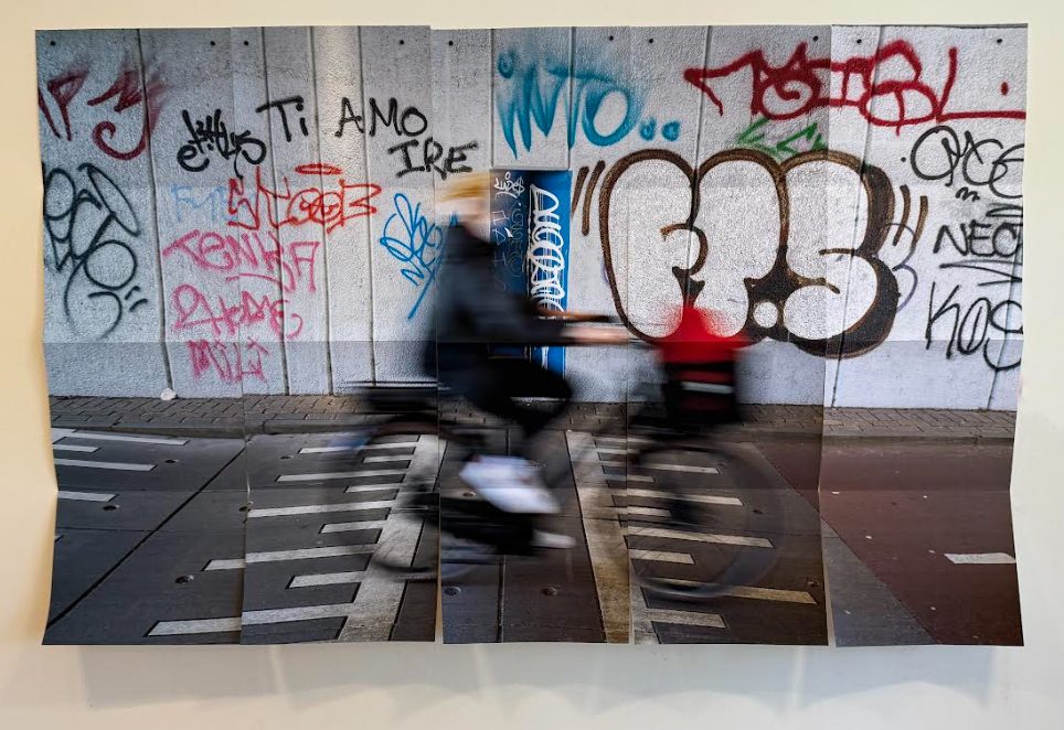 Printed photography of a motion-blurred cyclist riding past a wall of graffiti. The print has many fold marks.