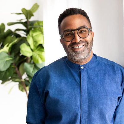 Dr. Jody Benjamin smiling at the camera in glasses and a purple button down shirt in front of a white background with foliage