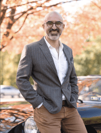 photo of a bald man with glasses in a suit jacket standing with hands in pockets.