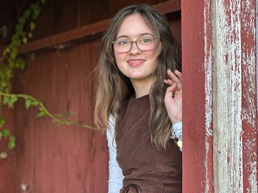 image of young woman with long hair and glasses standing near and old building with chipping red paint