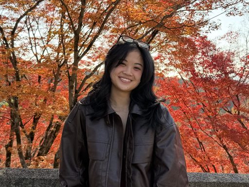Smiling person in a brown jacket stands against a backdrop of vivid autumn leaves.
