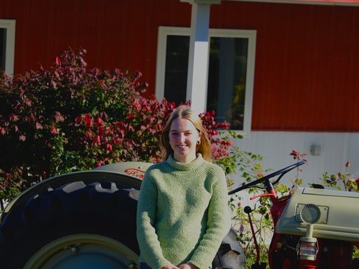 image of woman with long hair wearing a green sweater and jeans standing in front of an ole tractor