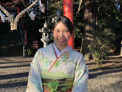 A woman wearing a light-colored, floral kimono smiles warmly in the sunlight. A red wooden pillar and stone lantern are behind her.