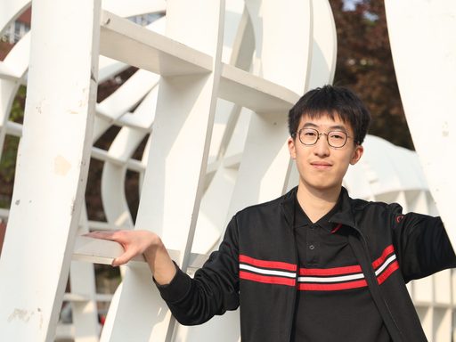 Young man in glasses and a black jacket with red stripes leans casually on a white geometric sculpture