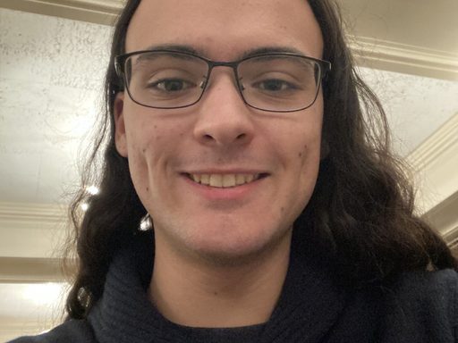 Young person with long hair and glasses smiling warmly, wearing a black sweater.
