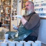 Ceramicist Ehren Tool discusses his work. Seated at a potter's wheel, Tool talks and gestures with his hands. A dozen newly made clay cups sit on a low table beside him.