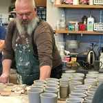 Ceramic Artist Ehren Tool prepares to begin decorating cups. Tool stands at a table next to a couple dozen newly made cups, with decorating tools and stamps on the table in front of him.
