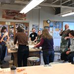 Ceramicist Ehren Tool speaks to students as they begin to decorate the cups. The students stand around a table filled with a couple dozen cups, with Tool standing behind them.