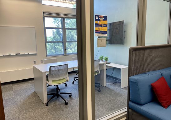Empty huddle space photographed from the outside showing table, whiteboard, and TV that students can use