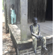 Statues of two boys in overalls. One is sitting on the edge of a wooden porch and the other is standing against the back wall of the porch. Statues by Woodrow Nash.