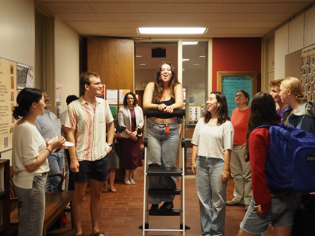 History senior Sophie Durdag '25 in jeans and a black tank top standing on a utility ladder addressing the AppleFest students.