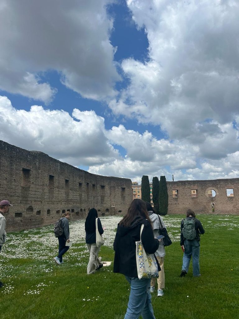 A sky full of clouds with Roman architecture and a group of students walking across a grass covered courtyard