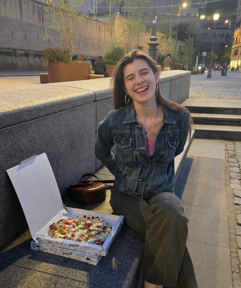 Victoria Semmelhack '25 laughing while sitting on a stone bench next to take away pizza.