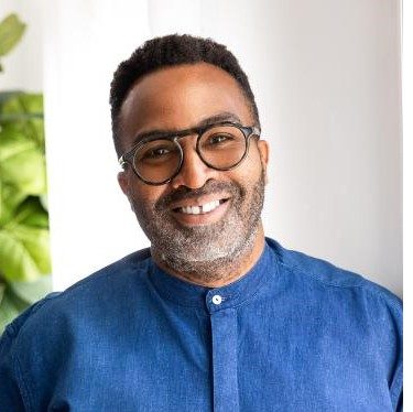 Headshot of Dr. Benjamin wearing a purple mandarin collar shirt in front of a white background and a green plant 