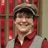 Sarah Ravely '04
Smiling directly at the camera, wearing a driver's cap and wire rimmed glasses red collared shirt and grey vest, standing in front of a red block window