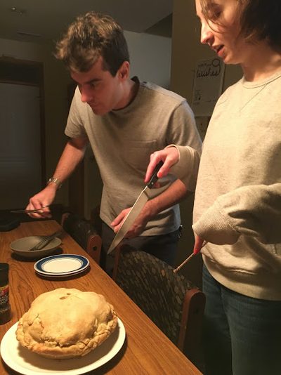 a handsome turtle-neck-wearing man holds a fork looking seriously at an apple pie while someone else holds a knife pointed at the pie