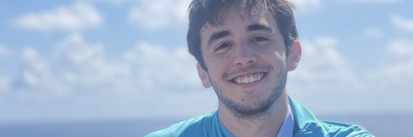 man with brown hair smiling near a body of water