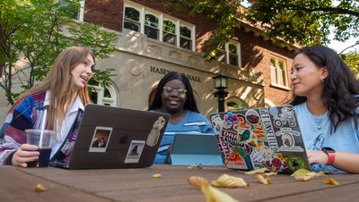 Three students sitting with their computers out on a picnic table outside Hasenstab Hall during the fall.