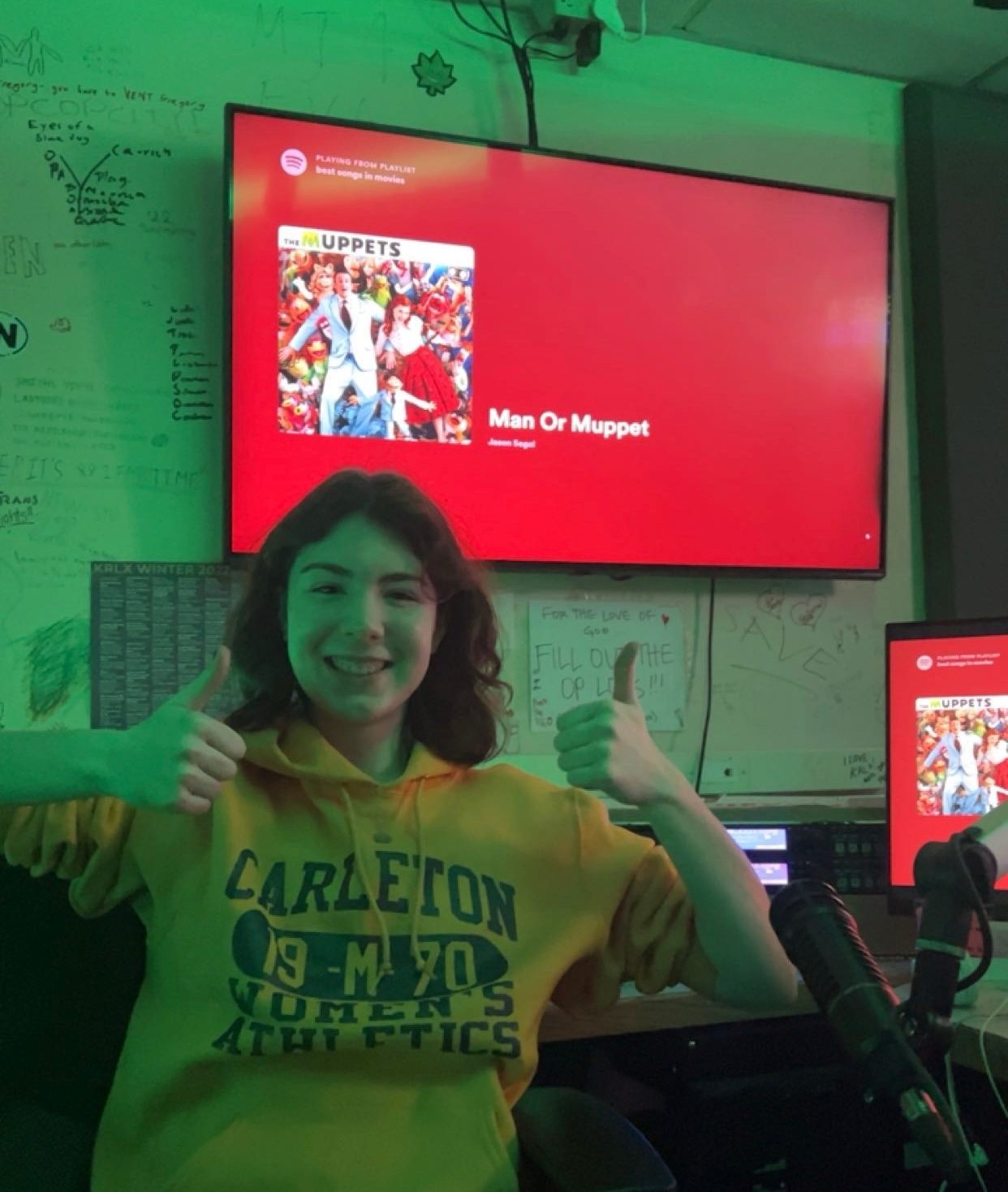 Girl in yellow sweatshirt giving a thumbs up with am I a Man or a Muppet playing on the screen behind her.
