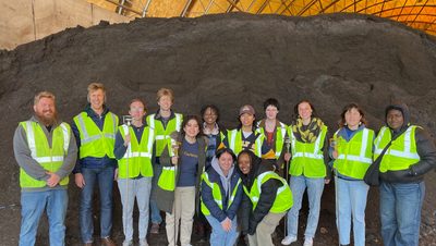 A group of students wearing yellow construction vests posed in front of dirt pile