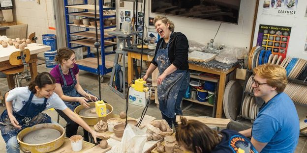 a laughing art professor stands above four smiling students making bowls on potter's wheels