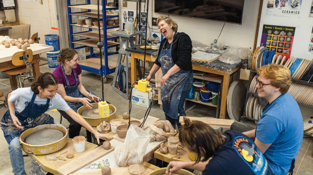a laughing art professor stands above four smiling students making bowls on potter's wheels
