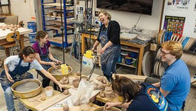 a laughing art professor stands above four smiling students making bowls on potter's wheels