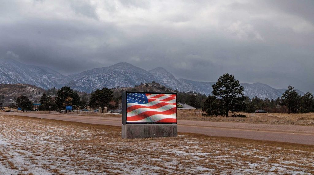 a digital american flag on a tv-billboard surrounded by snow, grass, and mountains.