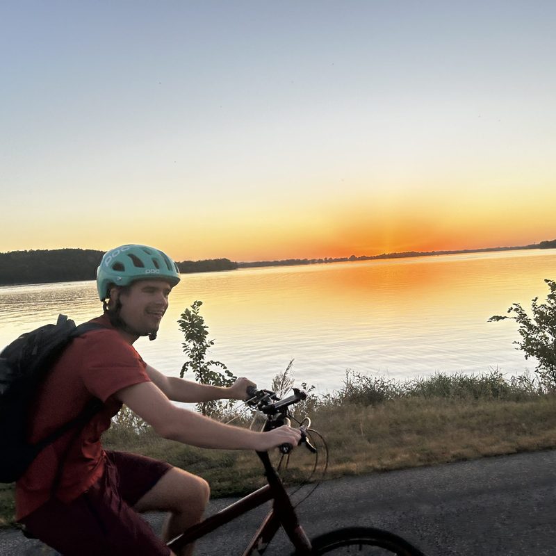 A student in a helmet bikes by a lake at sunset.
