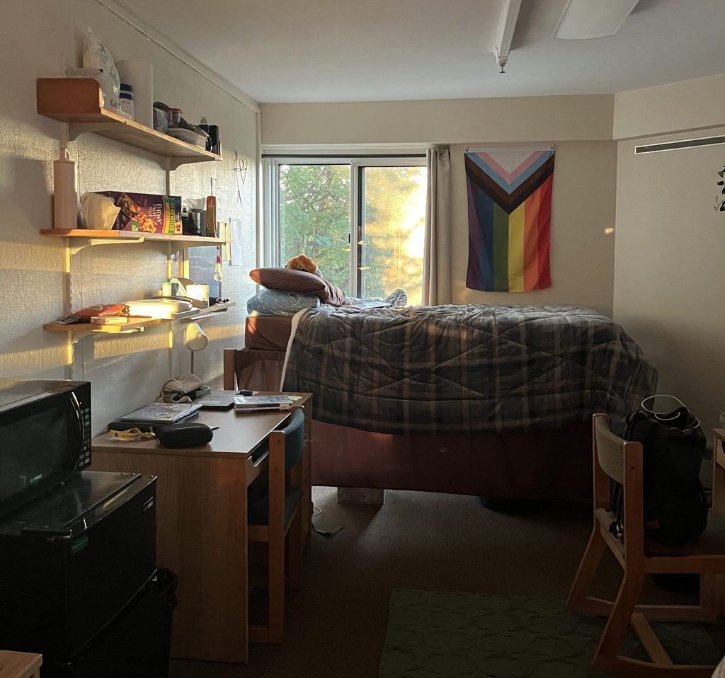A student's dorm room showing a bed, desk and mini-fridge with a microwave on top. 
