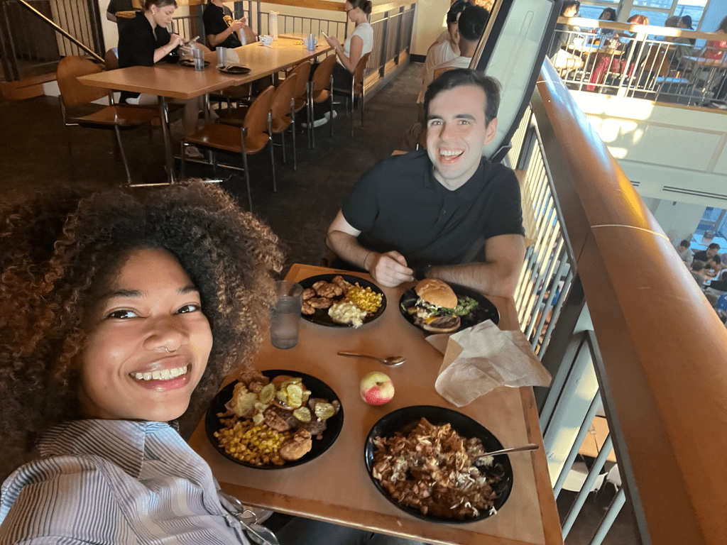Two students smile in a selfie with plates of food on the table