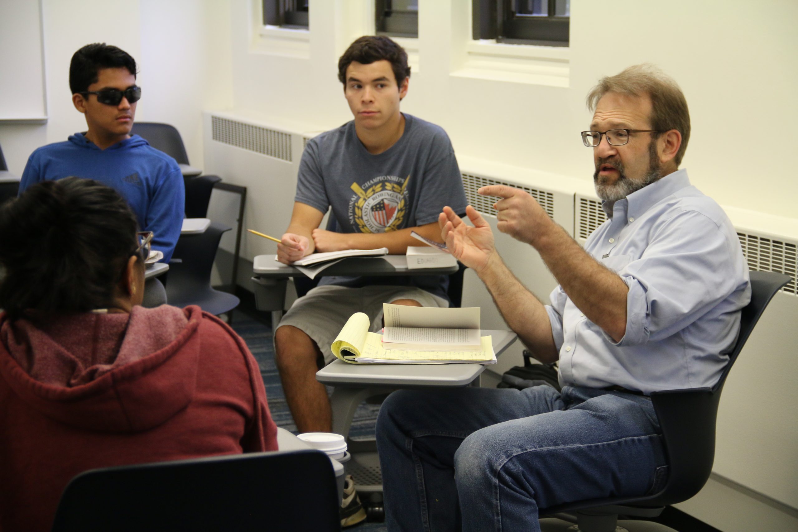 Professor Bill North and three students sit at desks in a circle while the students listen to Bill explaining some text