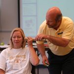 An AP Teacher sits in a chair with eyes closed while the consultant bends down to hold a paper towel tube to her ear.