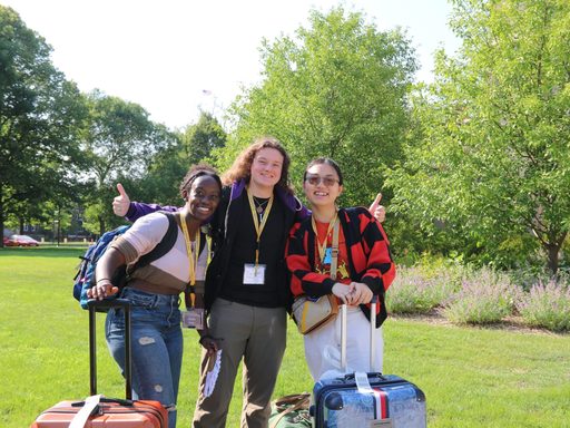 Three high school students smile with their suitcases on a lawn.
