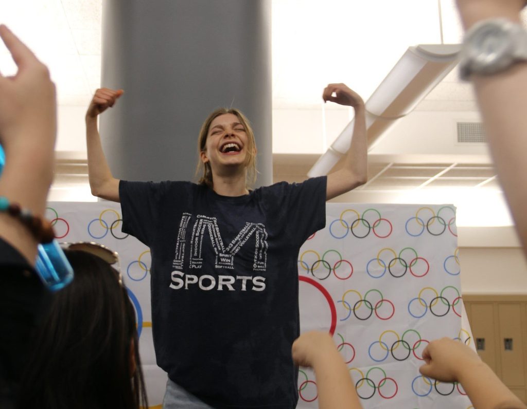 A student triumphantly raises her arms up in a sign of victory in front of an olympics-themed banner.