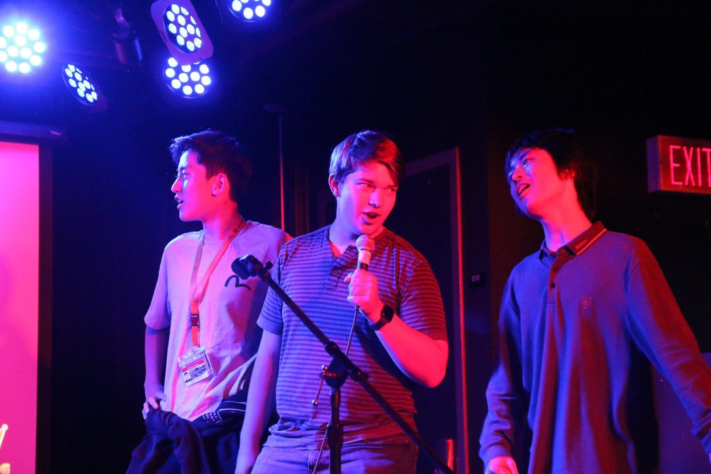 Three students sing Karaoke under the glow of purple stage lights.