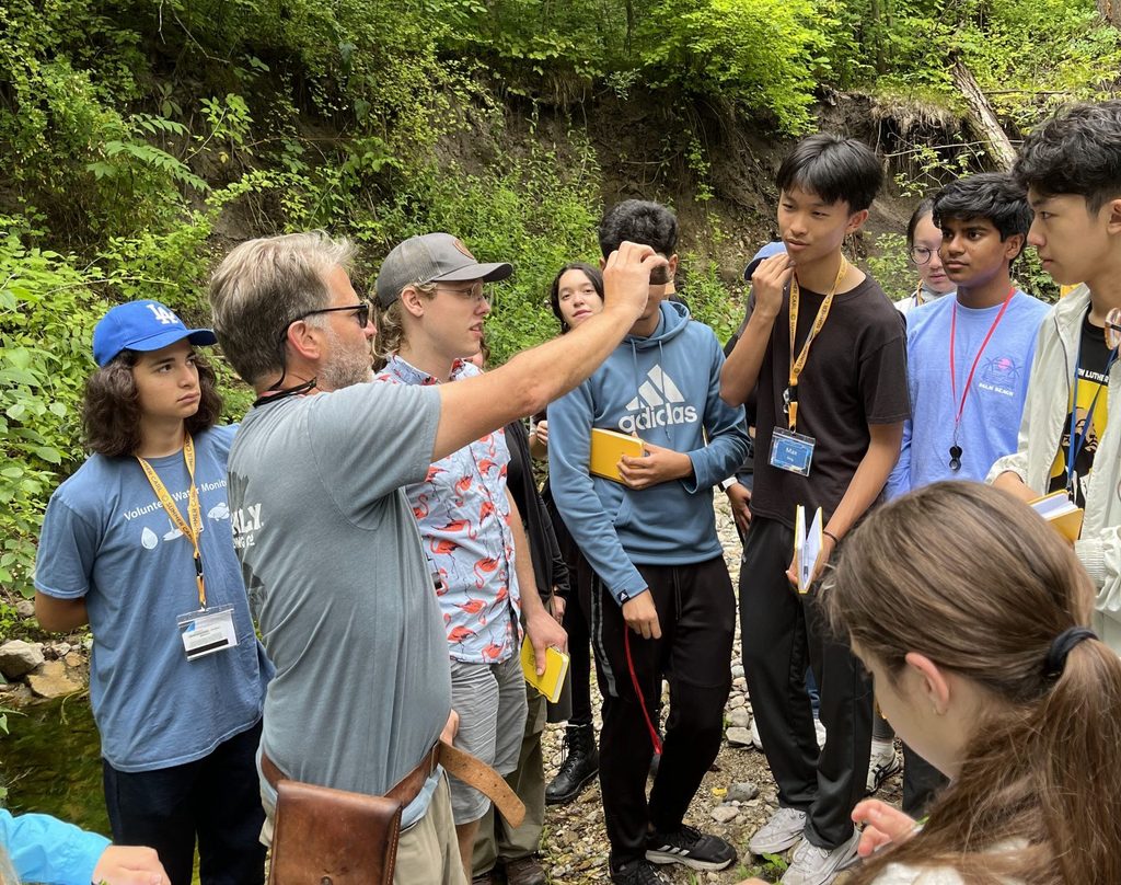 A group of students circle around a professor in the woods as he shows them a rock.