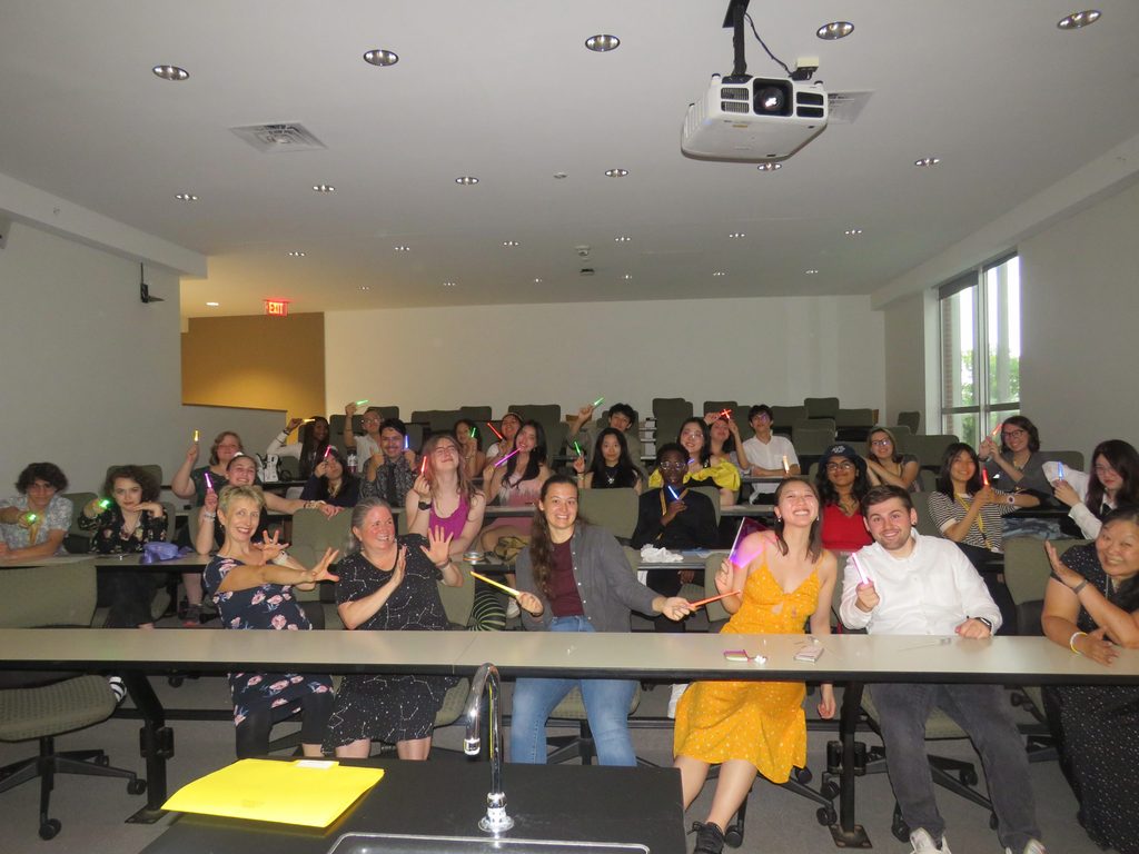 A large group of students and professors in a darkened classroom, each holding a glowing mini light saber.