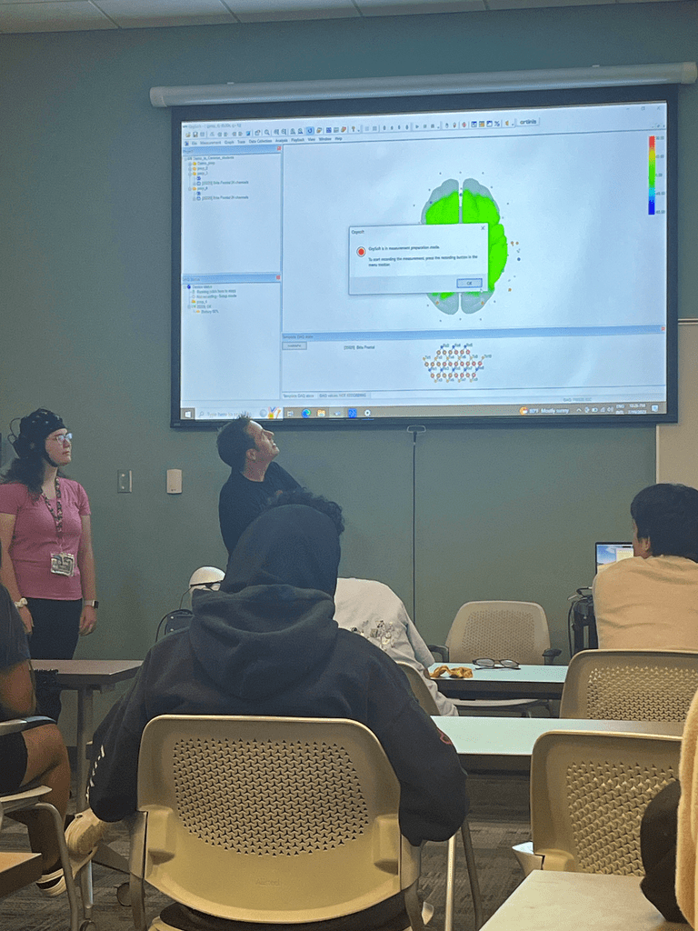 A student and professor stand at the front of the classroom, the professor is gesturing to the projector screen that shows a green schematic of the brain. 