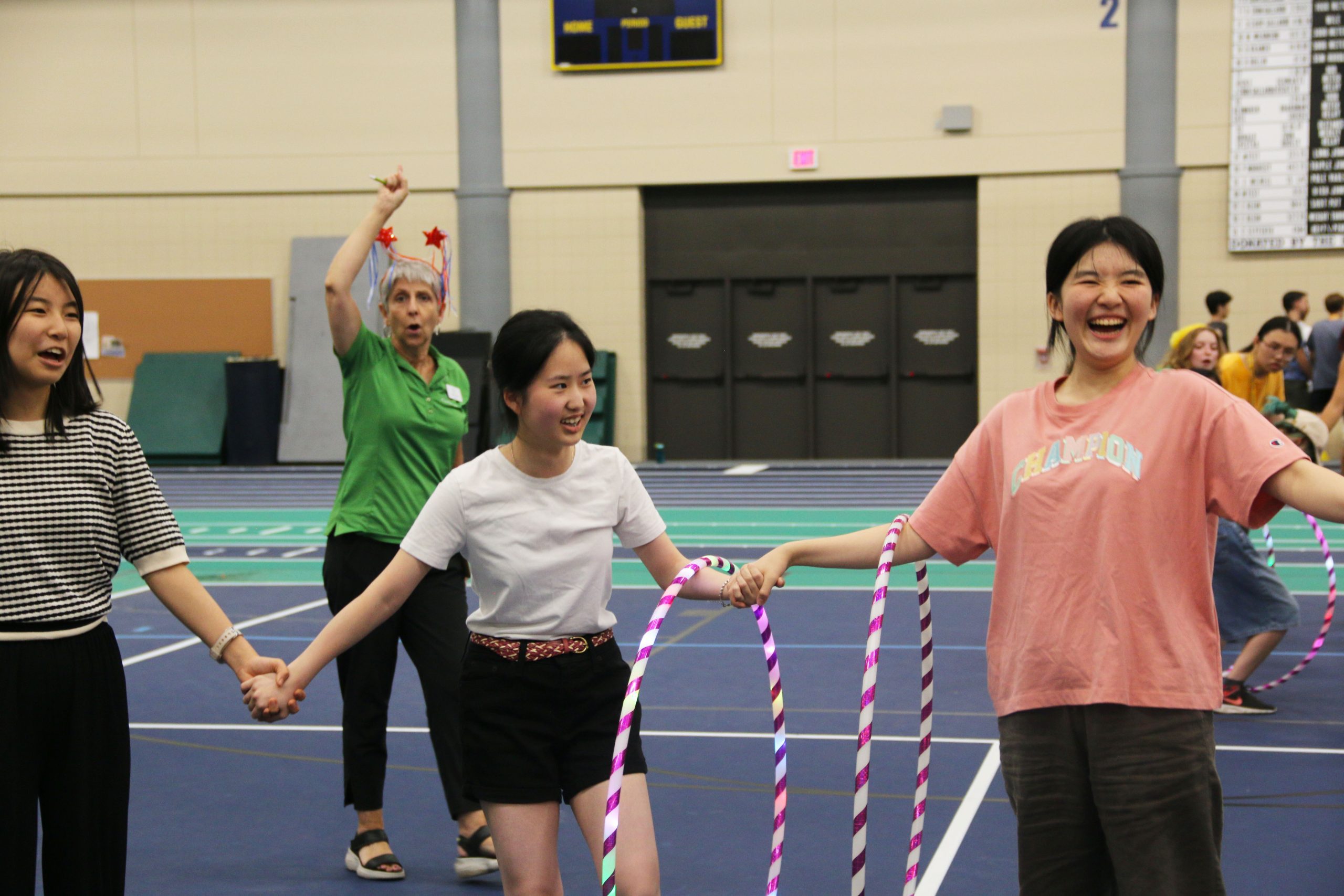 Three laughing students grasp hands with hula hoops hanging on their arms.