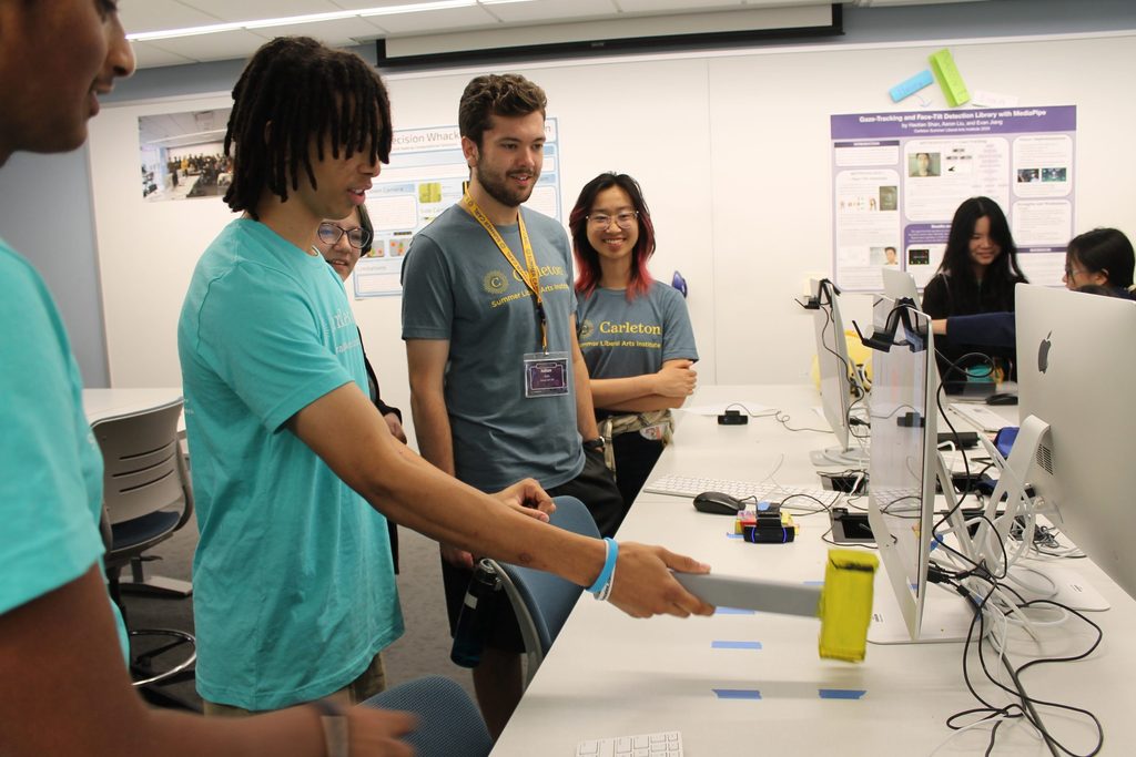 A student holding a mallet stands infront of a computer screen to play a computer science game while other students look on smiling.