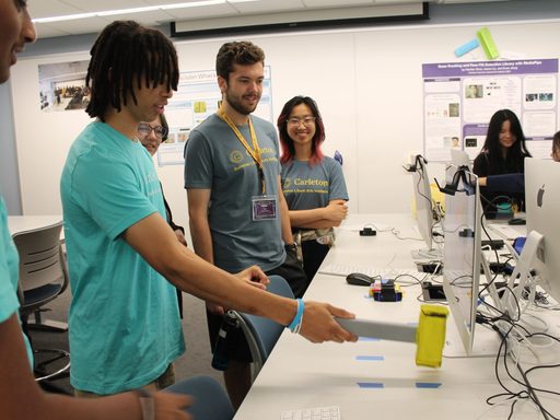 A student holding a mallet stands infront of a computer screen to play a computer science game while other students look on smiling.