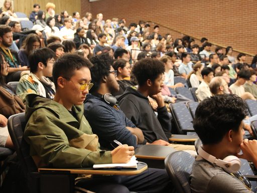 A student takes notes in a notebook while sitting in a tiered auditorium filled with students listening to a speaker.