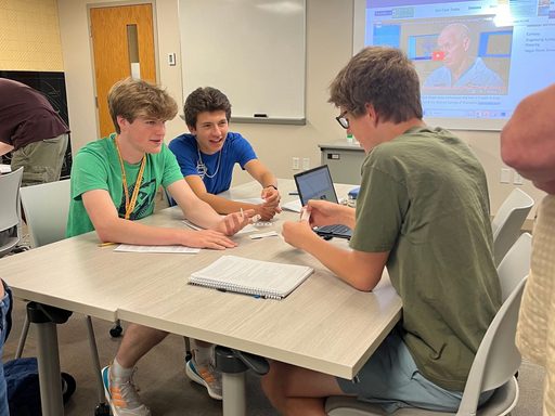 Three students sit at a classroom table holding a small group discussion.