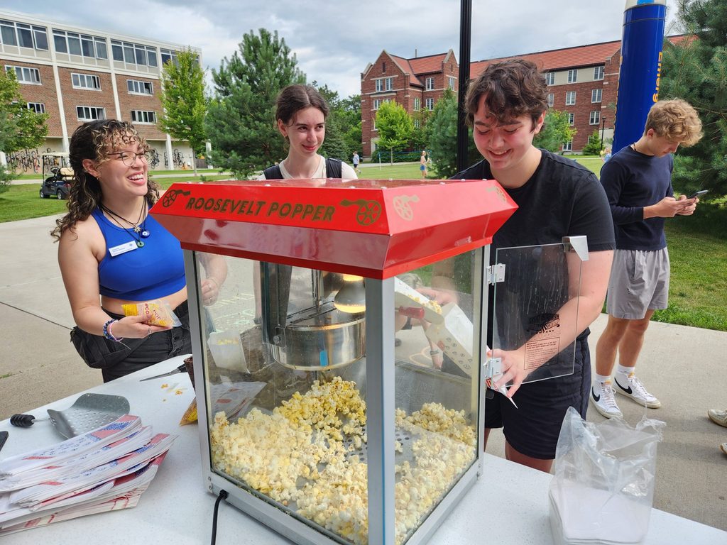 Three student workers smile while standing behind a popcorn machine near a green space on campus.