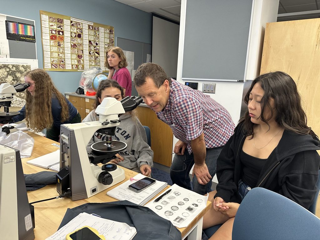 A professor leans into a microscope to assist students in their assignment while students wathc